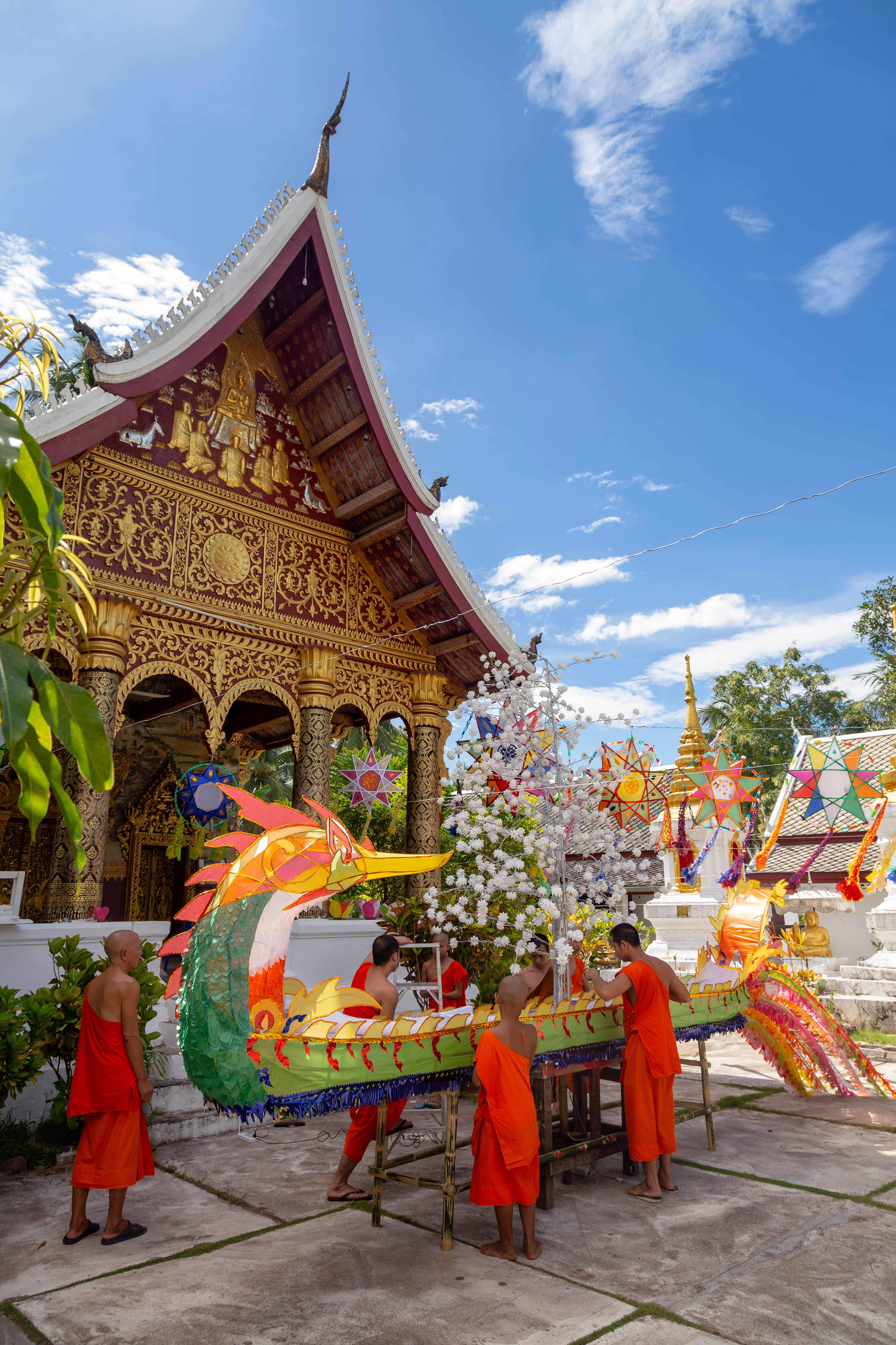 Buddhist monks in orange robes assemble a large decorative naga (serpent) boat float outside a temple in Luang Prabang, Laos, with colorful star-shaped lanterns hanging in the temple courtyard under a blue sky.