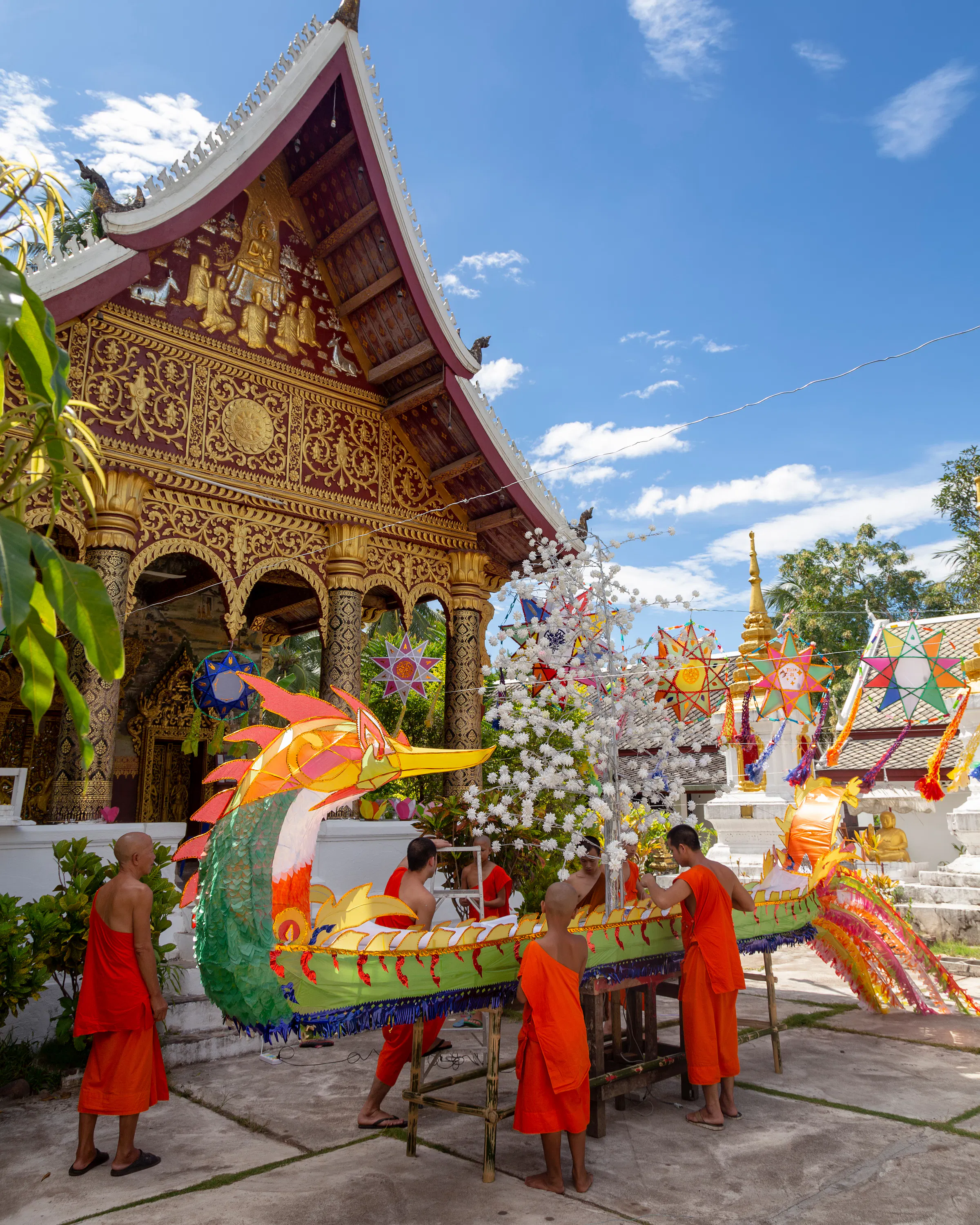 Buddhist monks in orange robes assemble a large decorative naga (serpent) boat float outside a temple in Luang Prabang, Laos, with colorful star-shaped lanterns hanging in the temple courtyard under a blue sky.