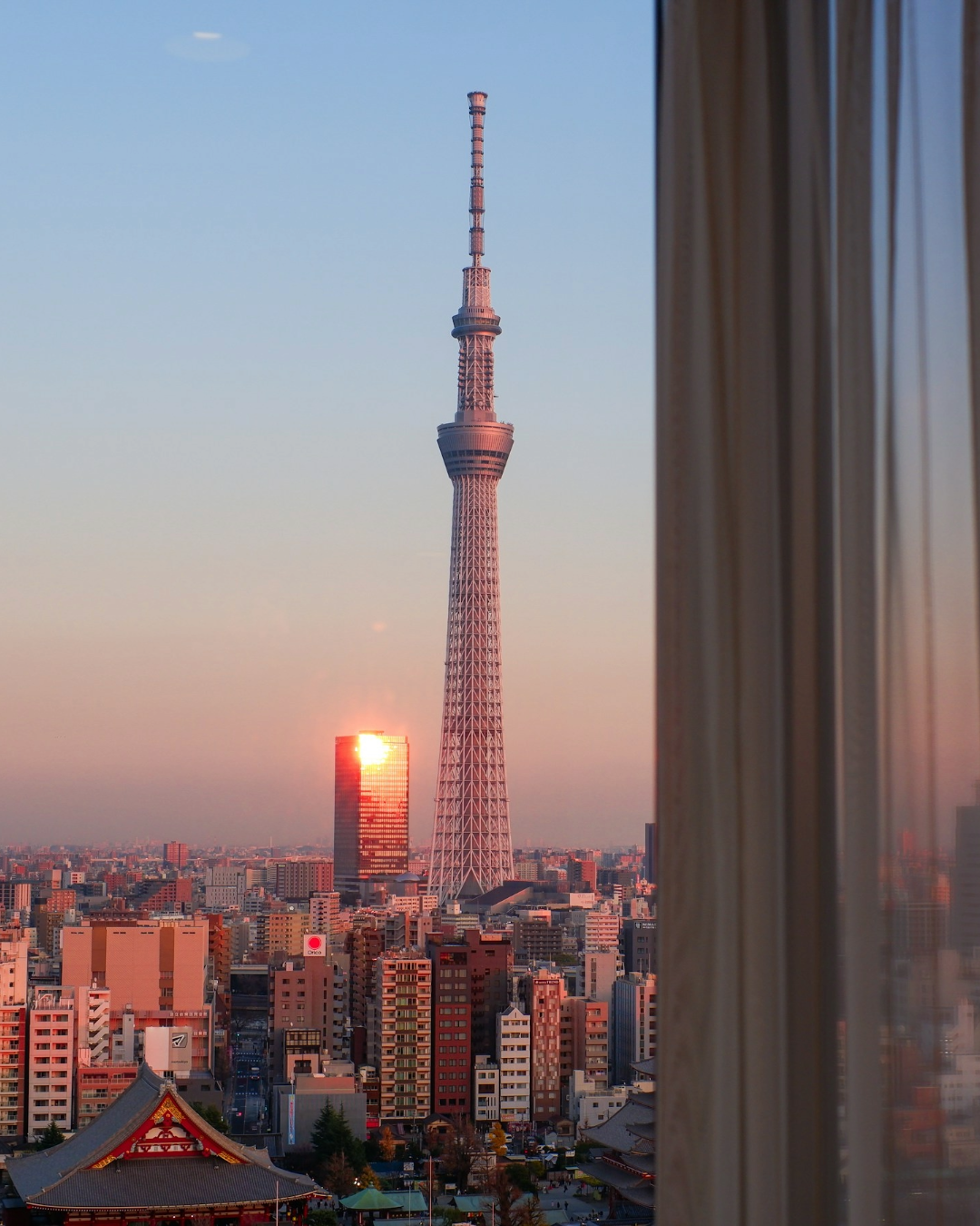 Tokyo Skytree rising above the city skyline at sunset, with the rooftops of Asakusa and a traditional temple in the foreground.
