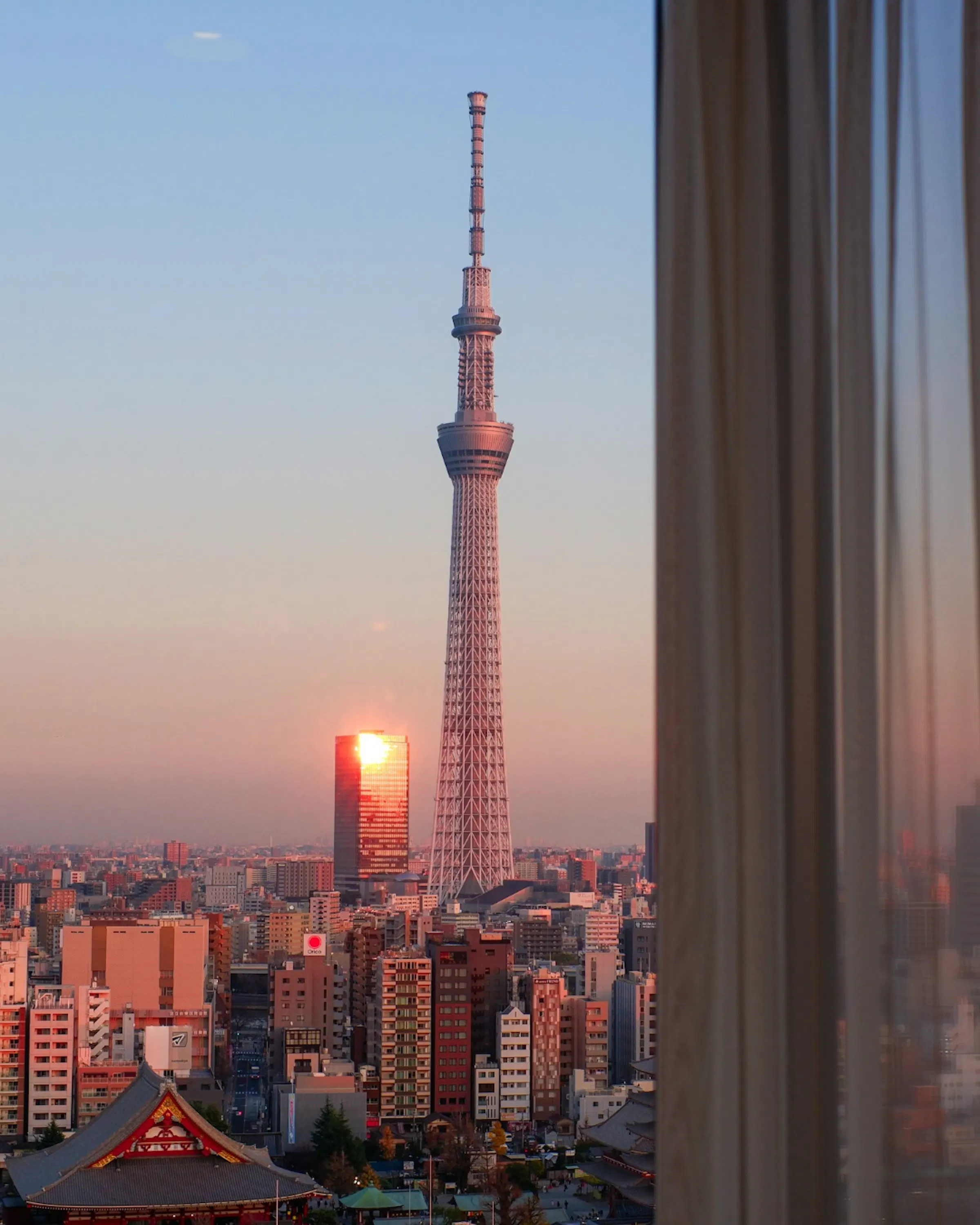 Tokyo Skytree rising above the city skyline at sunset, with the rooftops of Asakusa and a traditional temple in the foreground.