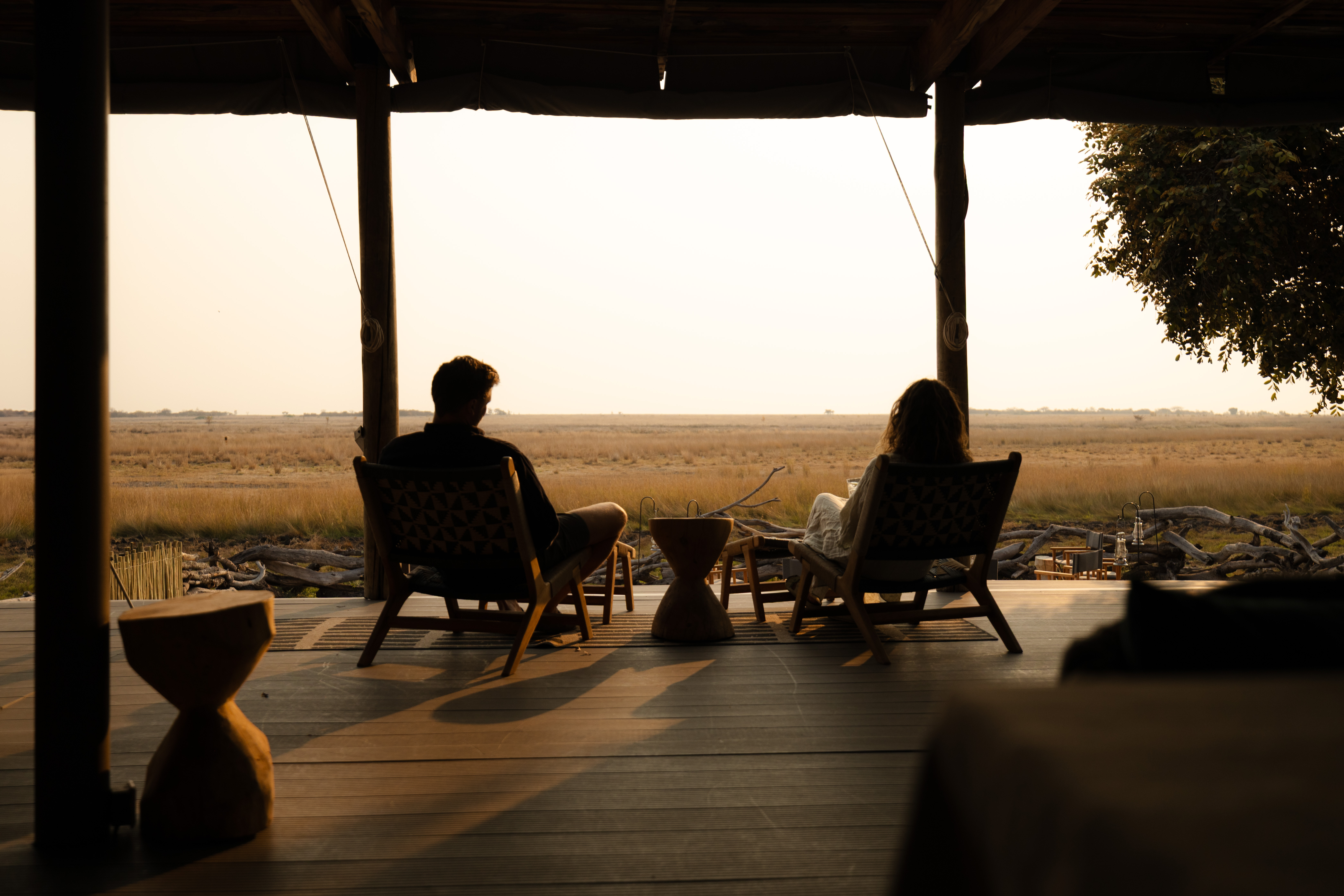 A couple sits in silhouette on a shaded lodge deck in Zambia, facing out over open golden grassland at sunset.