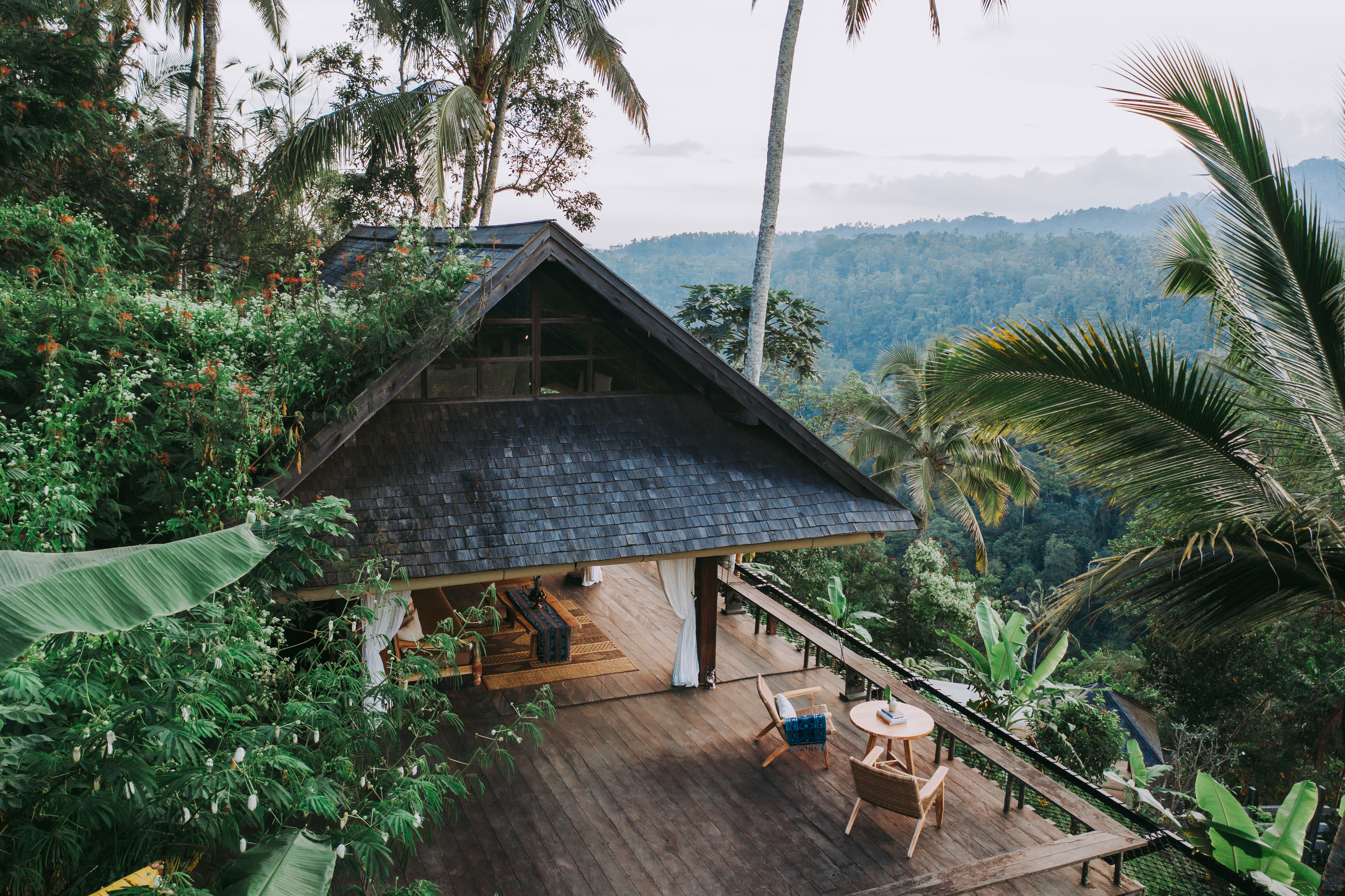 A shingled, peaked-roof villa with an open-air deck and lounge chairs sits perched among palm trees and tropical foliage, overlooking a misty, forested valley in Bali's Ubud highlands.