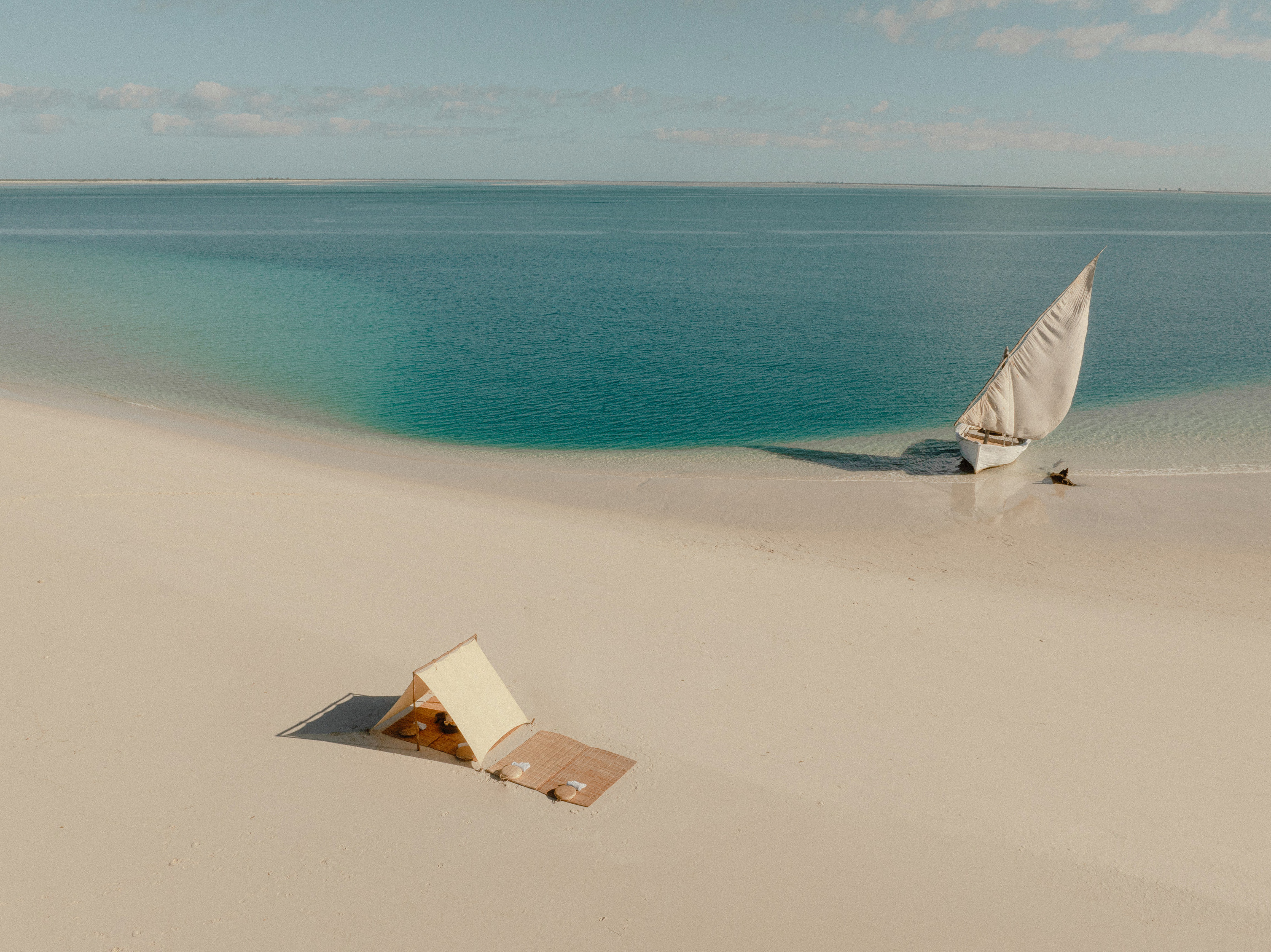 An aerial view of a private picnic tent set up on a sandbar in Mozambique, and a traditional dhow anchored just offshore in turquoise water.
