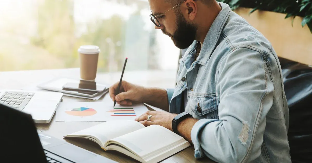 Male, graduate-aged student sitting at table with laptop, coffee and study papers