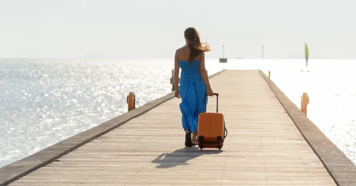 Woman in blue dress walking on a pier traiuling a roller suitcase