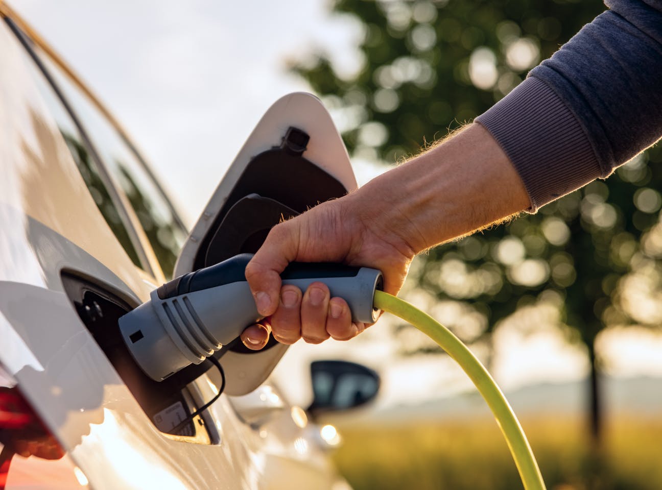 Un primo piano mostra una mano che inserisce un cavo di ricarica in un’auto elettrica bianca parcheggiata all’aperto. Il connettore grigio con cavo giallo è collegato alla presa sul lato della vettura, con lo sportellino aperto. Sullo sfondo, leggermente sfocato, si intravedono alberi verdi e la luce calda del tramonto, che evocano un contesto naturale e sostenibile.