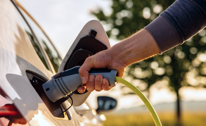 Un primo piano mostra una mano che inserisce un cavo di ricarica in un’auto elettrica bianca parcheggiata all’aperto. Il connettore grigio con cavo giallo è collegato alla presa sul lato della vettura, con lo sportellino aperto. Sullo sfondo, leggermente sfocato, si intravedono alberi verdi e la luce calda del tramonto, che evocano un contesto naturale e sostenibile.