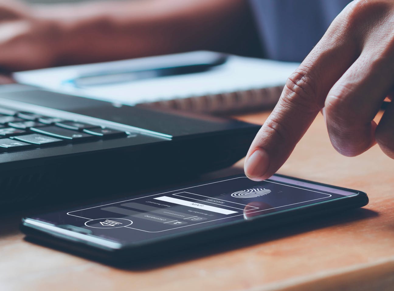 Dedo tocando la pantalla de un smartphone con interfaz de pago digital, junto a un portátil sobre un escritorio.