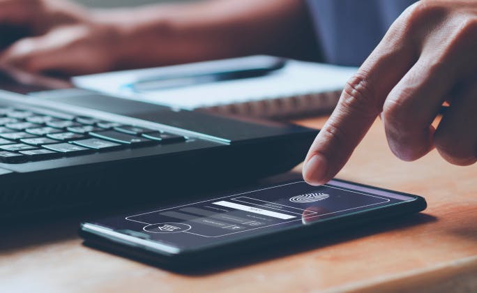 Dedo tocando la pantalla de un smartphone con interfaz de pago digital, junto a un portátil sobre un escritorio.