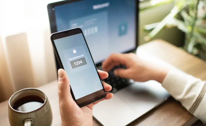 Persona alla scrivania che consulta sullo smartphone un codice di verifica per l’autenticazione a due fattori mentre usa il laptop.
