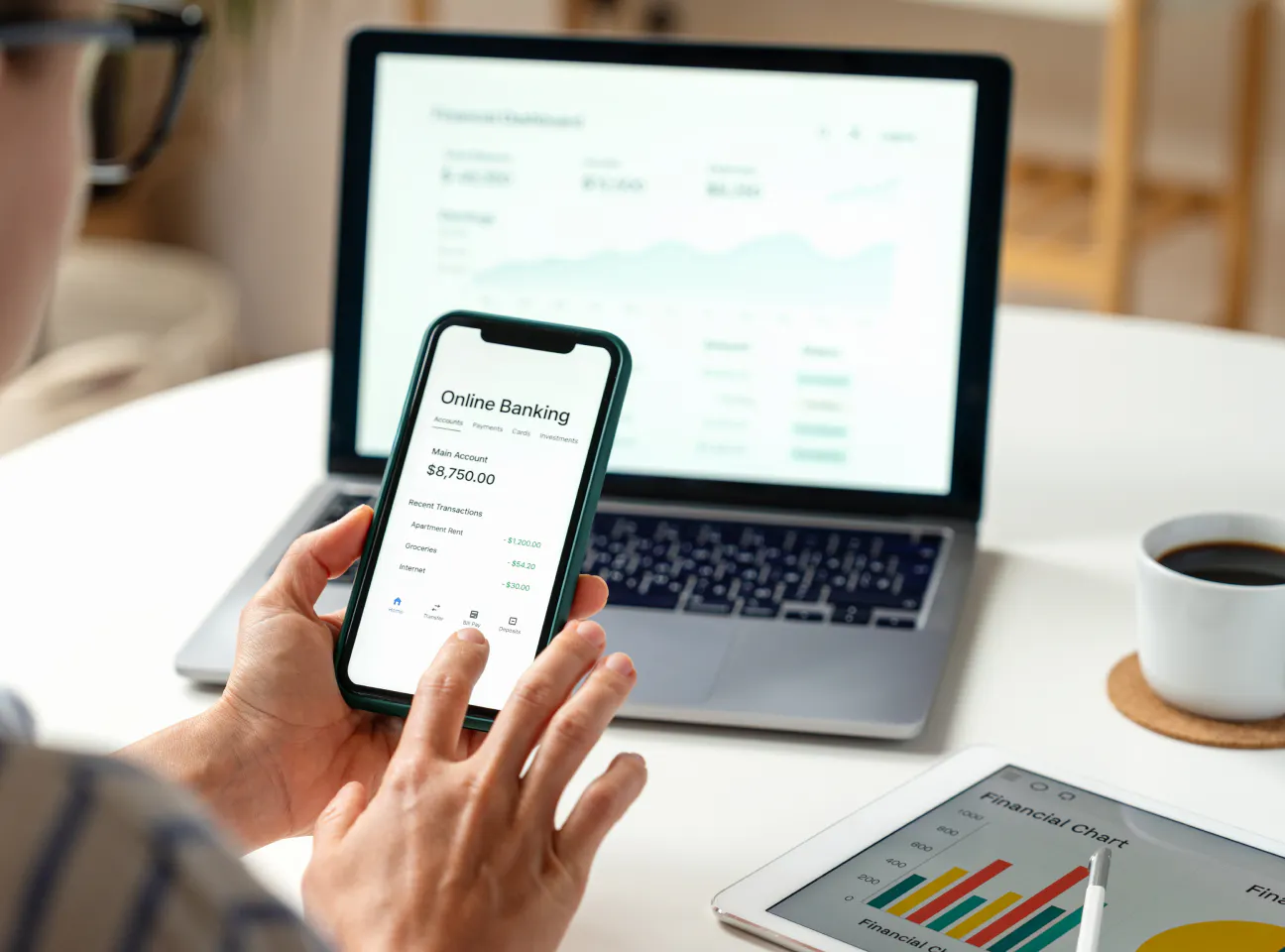 Persona che utilizza un’app di online banking su smartphone davanti a un laptop, con dashboard finanziaria sullo schermo; su un tavolo sono presenti anche una tazza di caffè e un tablet con grafico.