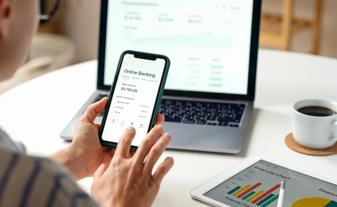 Persona che utilizza un’app di online banking su smartphone davanti a un laptop, con dashboard finanziaria sullo schermo; su un tavolo sono presenti anche una tazza di caffè e un tablet con grafico.