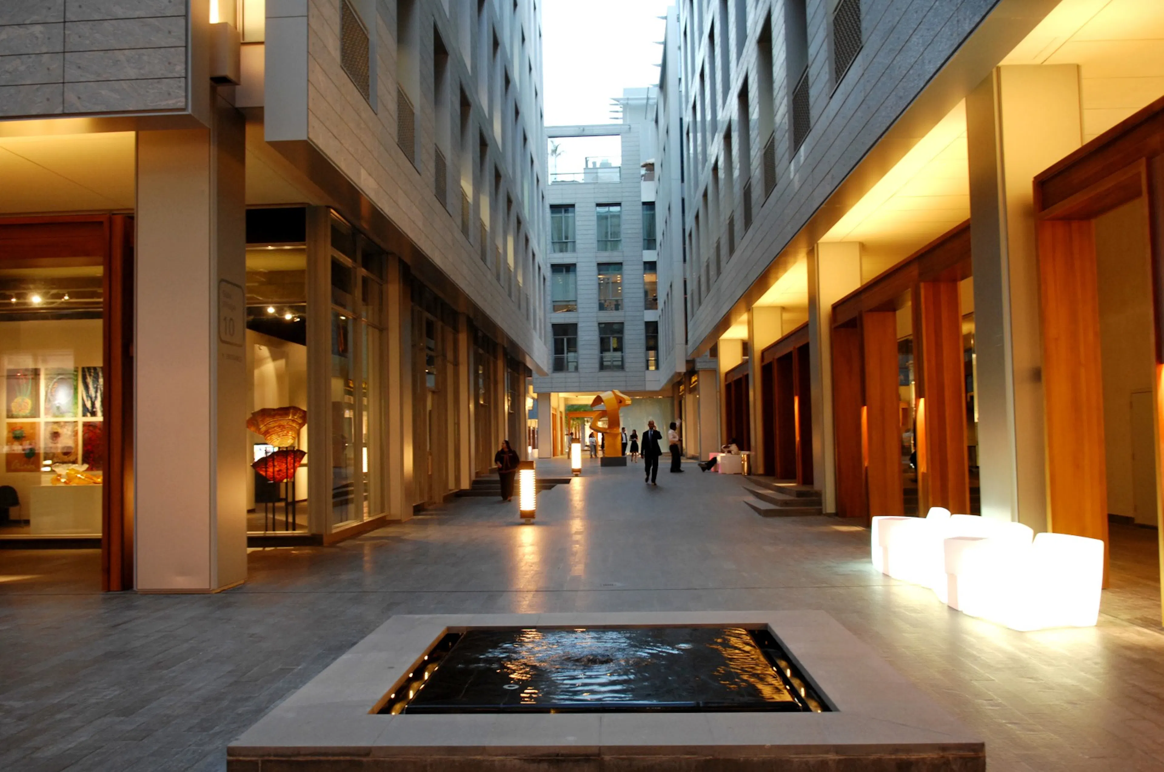 A modern architectural walkway lined with retail storefronts and a reflecting pool, framed by multi-storey buildings