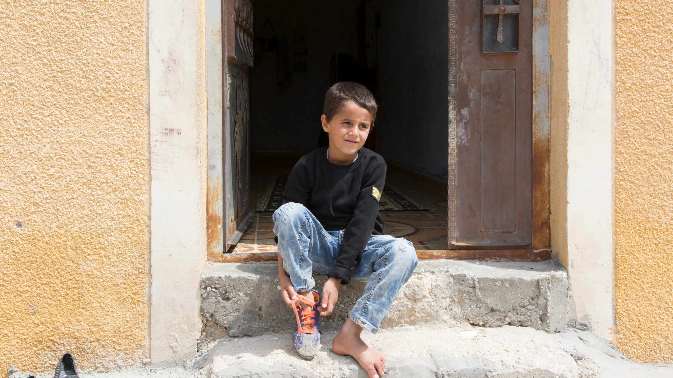 A young boy sitting in a doorway, tying his shoe, in a rural village setting