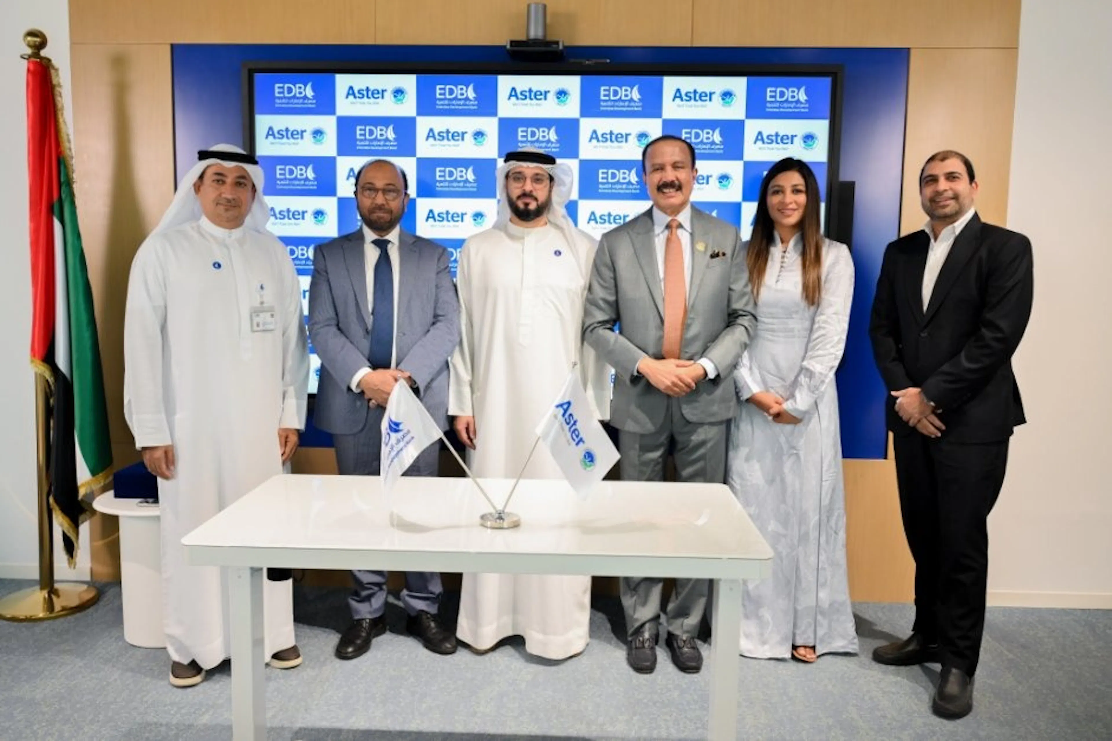 Six people standing at a signing-ceremony table in front of an Aster-branded backdrop, with a UAE flag alongside