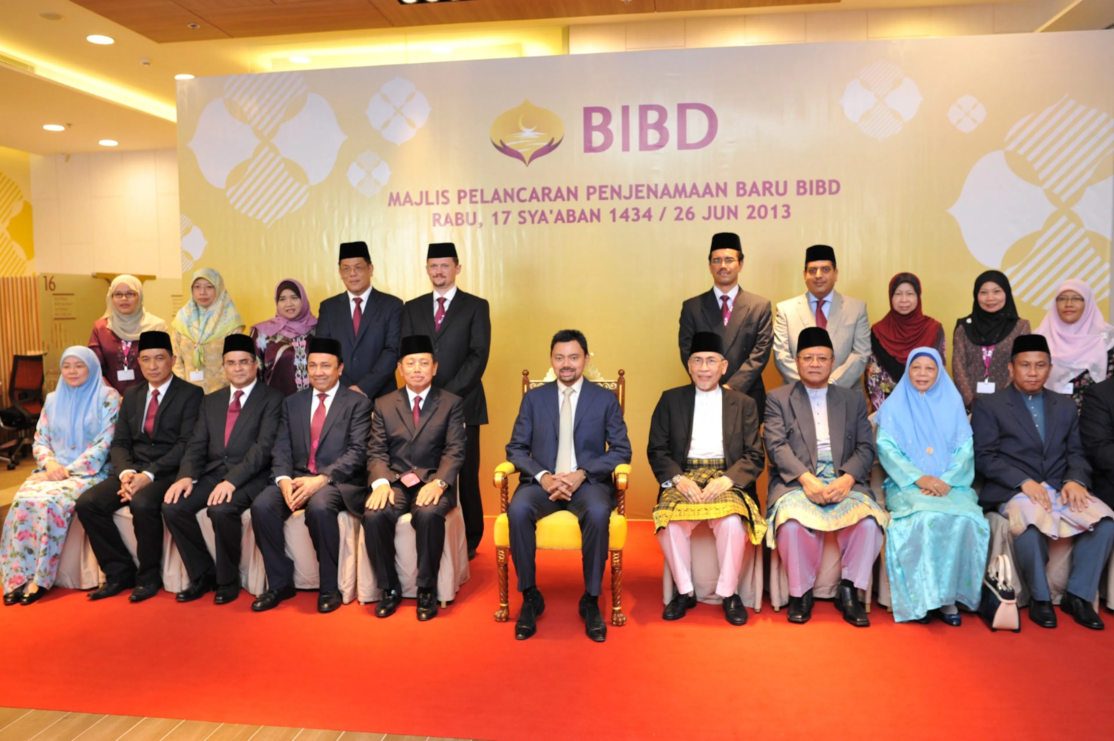 A formal group portrait on stage at a BIBD rebranding ceremony, attendees seated in traditional and formal dress against a decorated backdrop