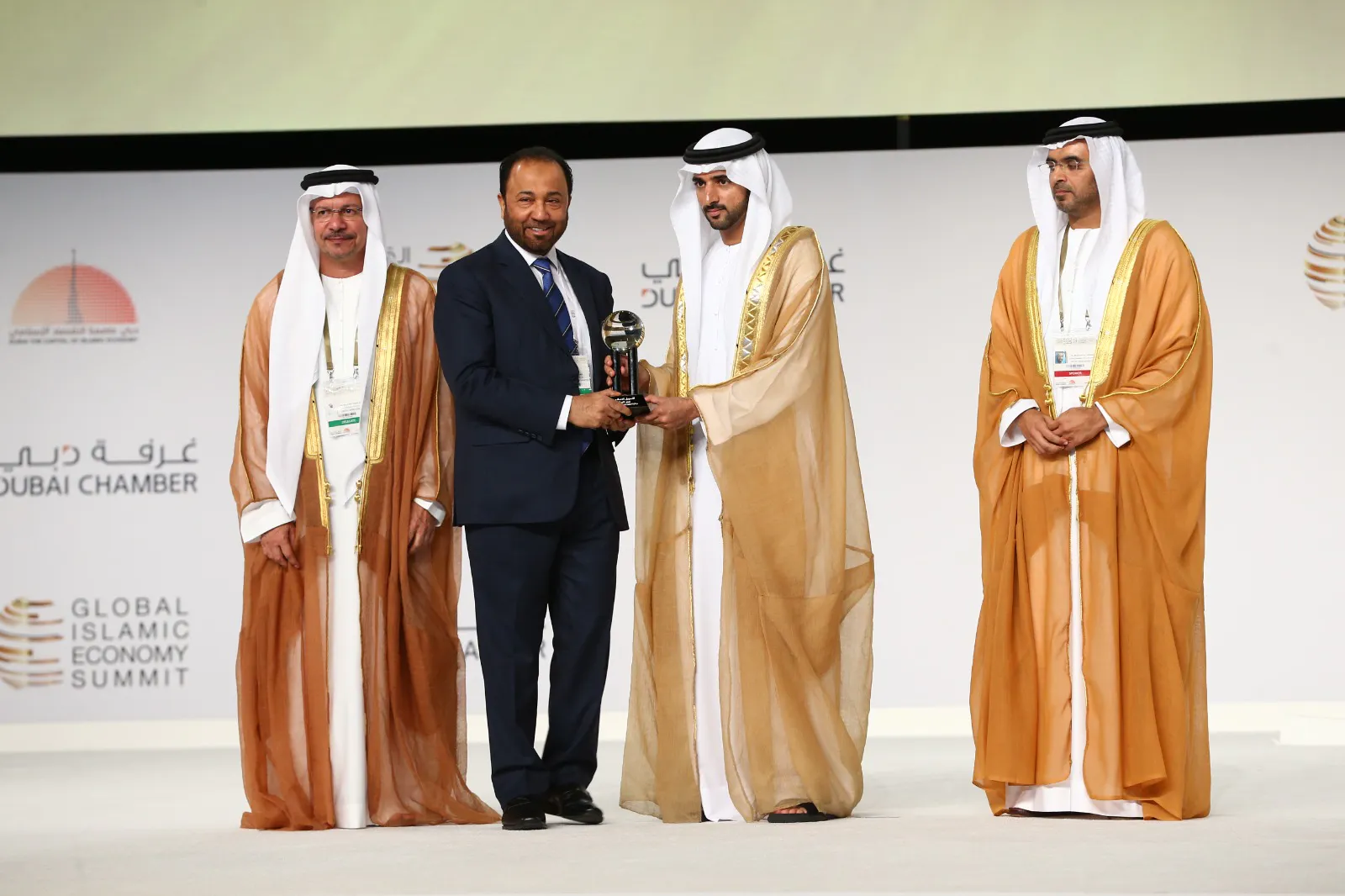 Three men in traditional Gulf dress on stage at the Global Islamic Economy Summit, one presenting an award to another in front of a Dubai Chamber backdrop