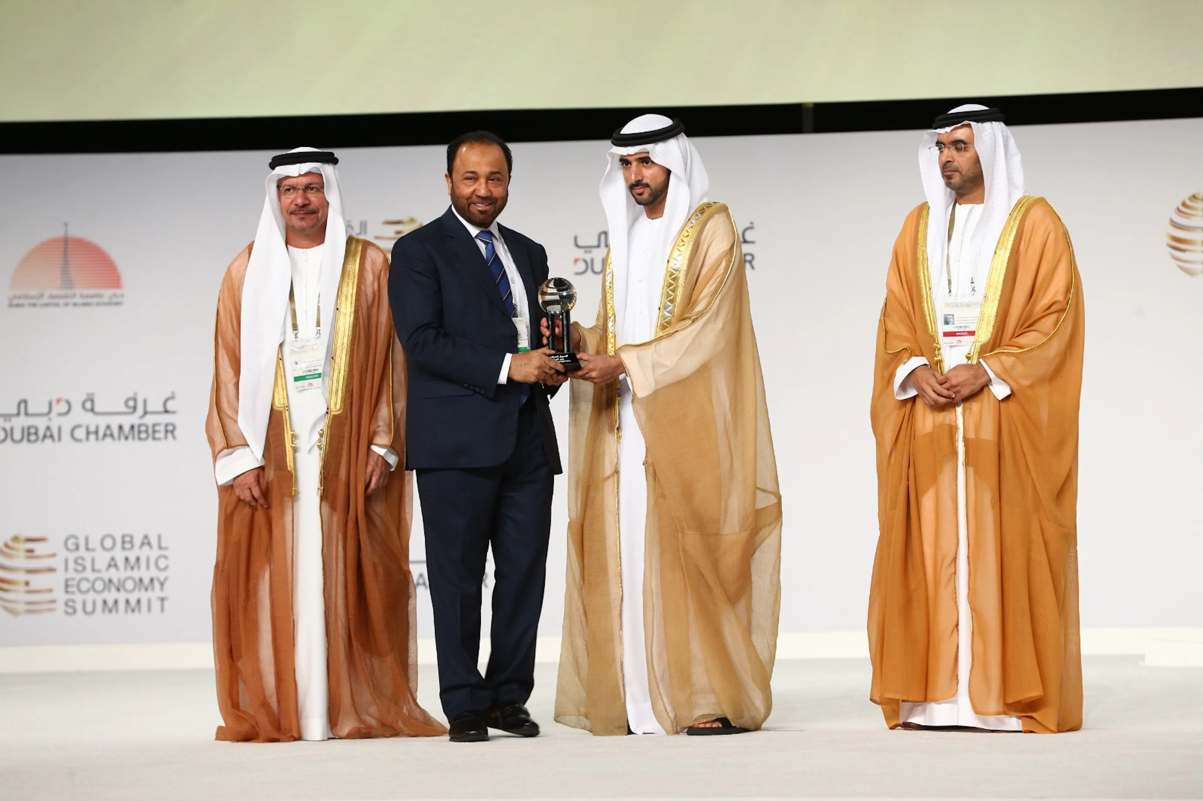 Three men in traditional Gulf dress on stage at the Global Islamic Economy Summit, one presenting an award to another in front of a Dubai Chamber backdrop
