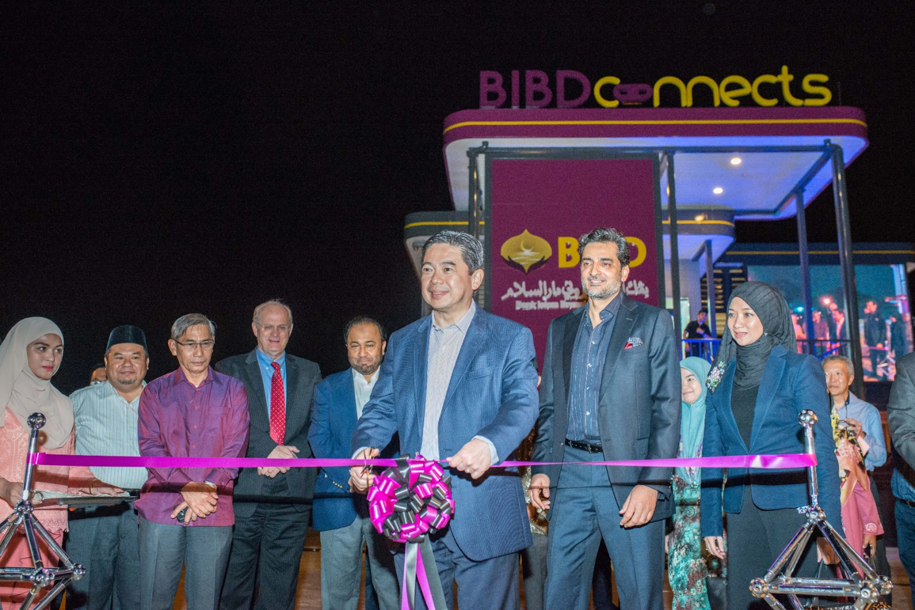 A group cutting a ribbon at the night-time opening of BIBD Connects, with the illuminated storefront lit up behind them