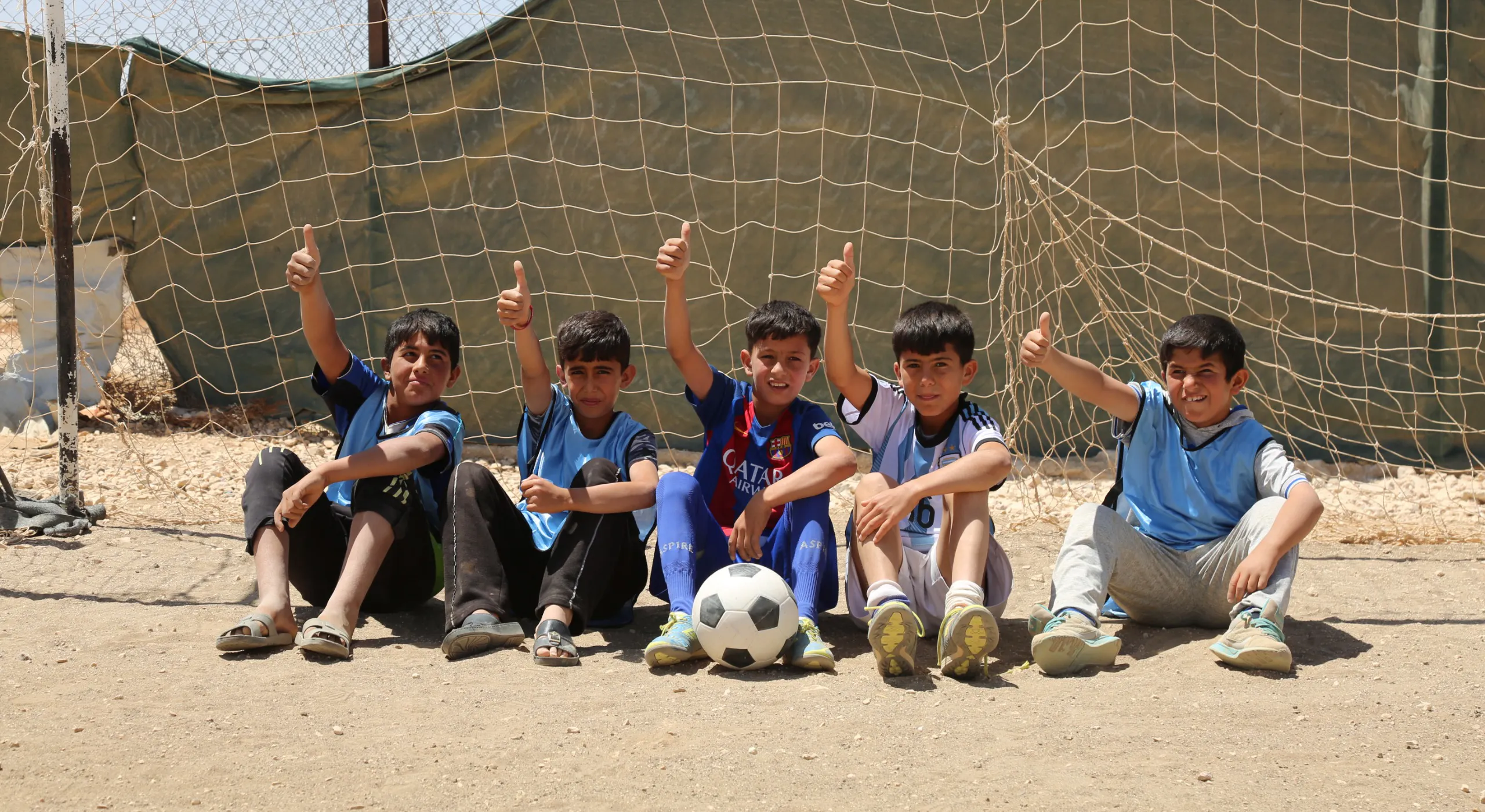 Five young boys sitting in a row against a soccer goal net, smiling and giving a thumbs up
