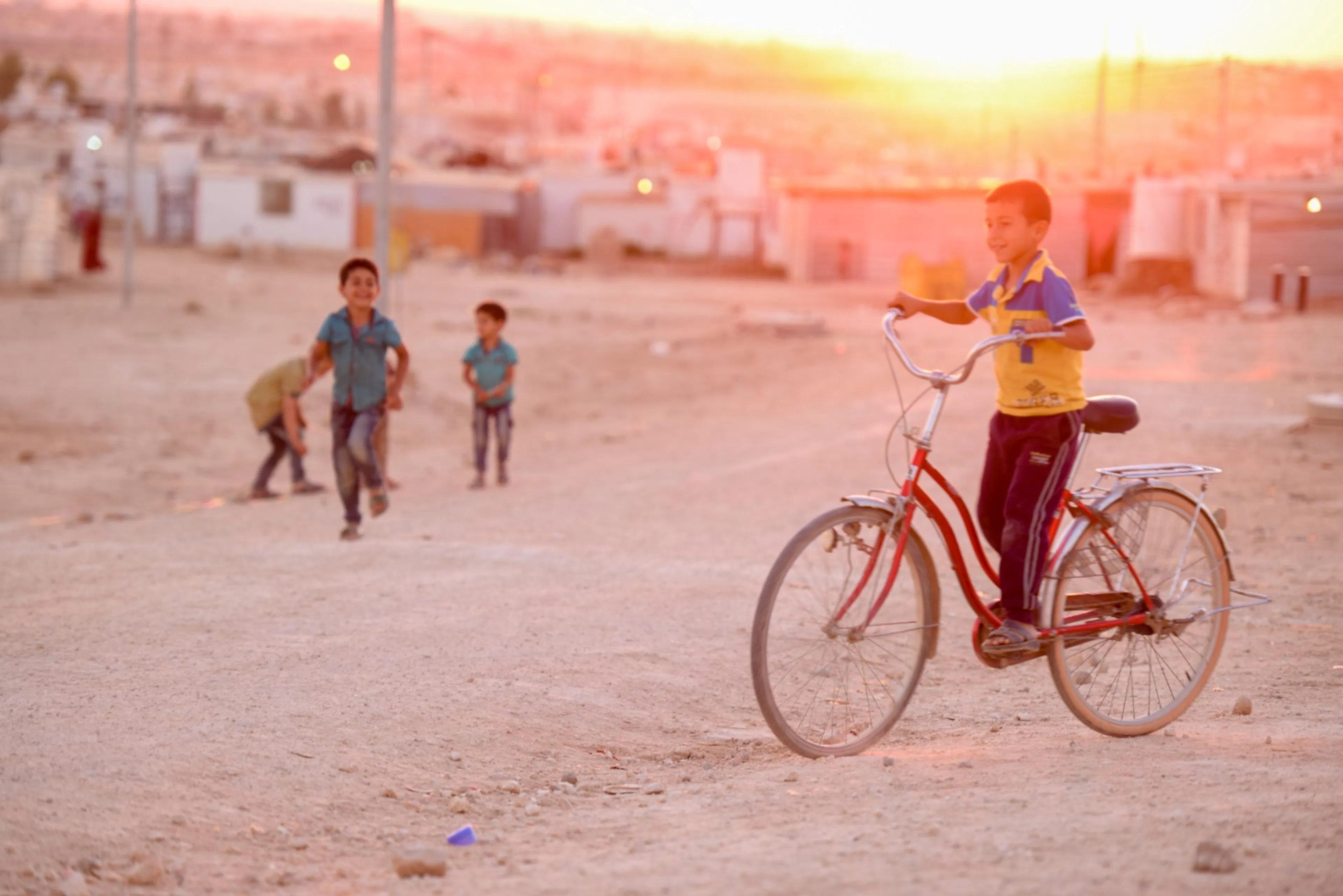 A boy riding a bicycle through a refugee camp at sunset, with other children playing in the background