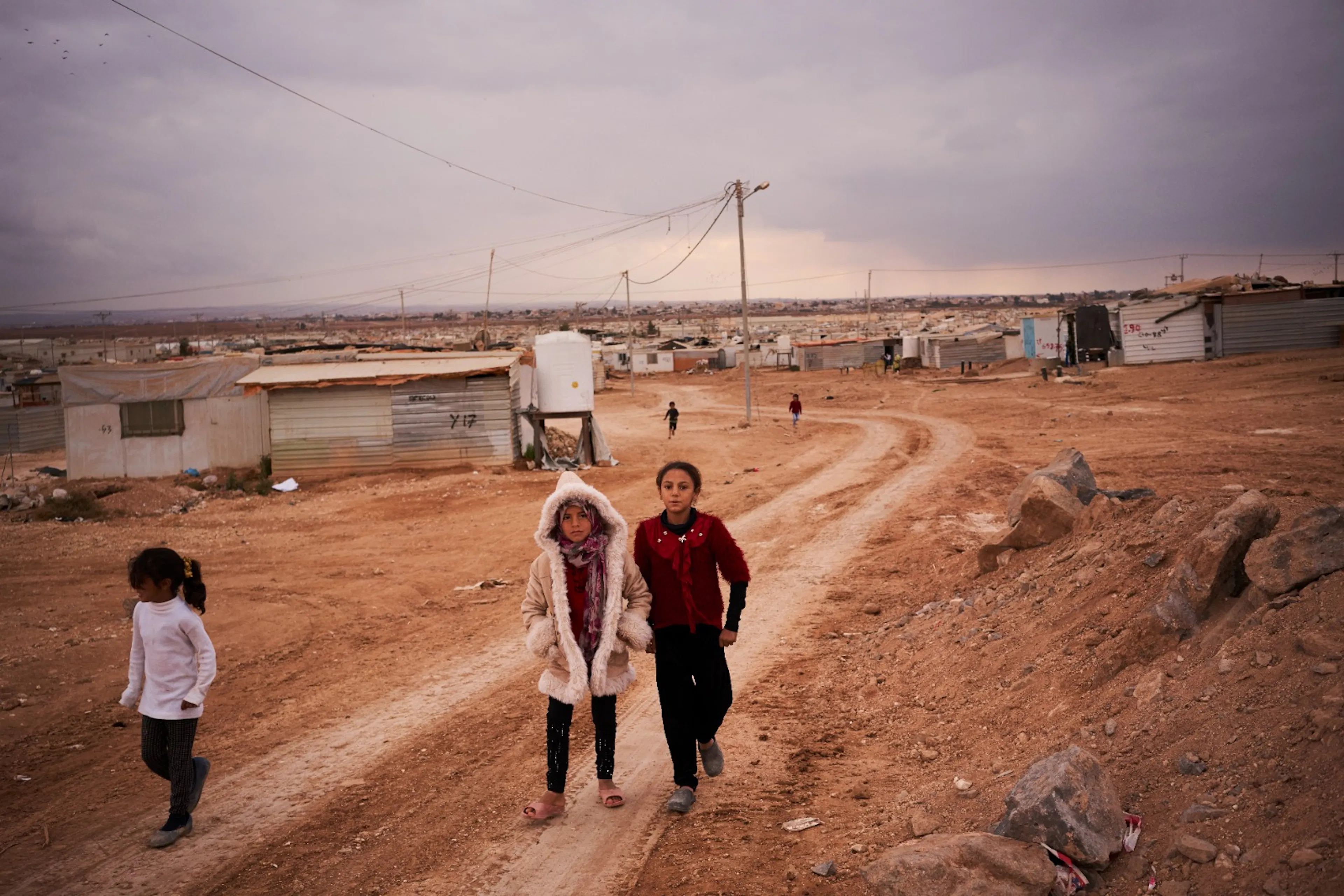 Two children walking along a dirt road through a refugee camp, with rows of shelters in the distance
