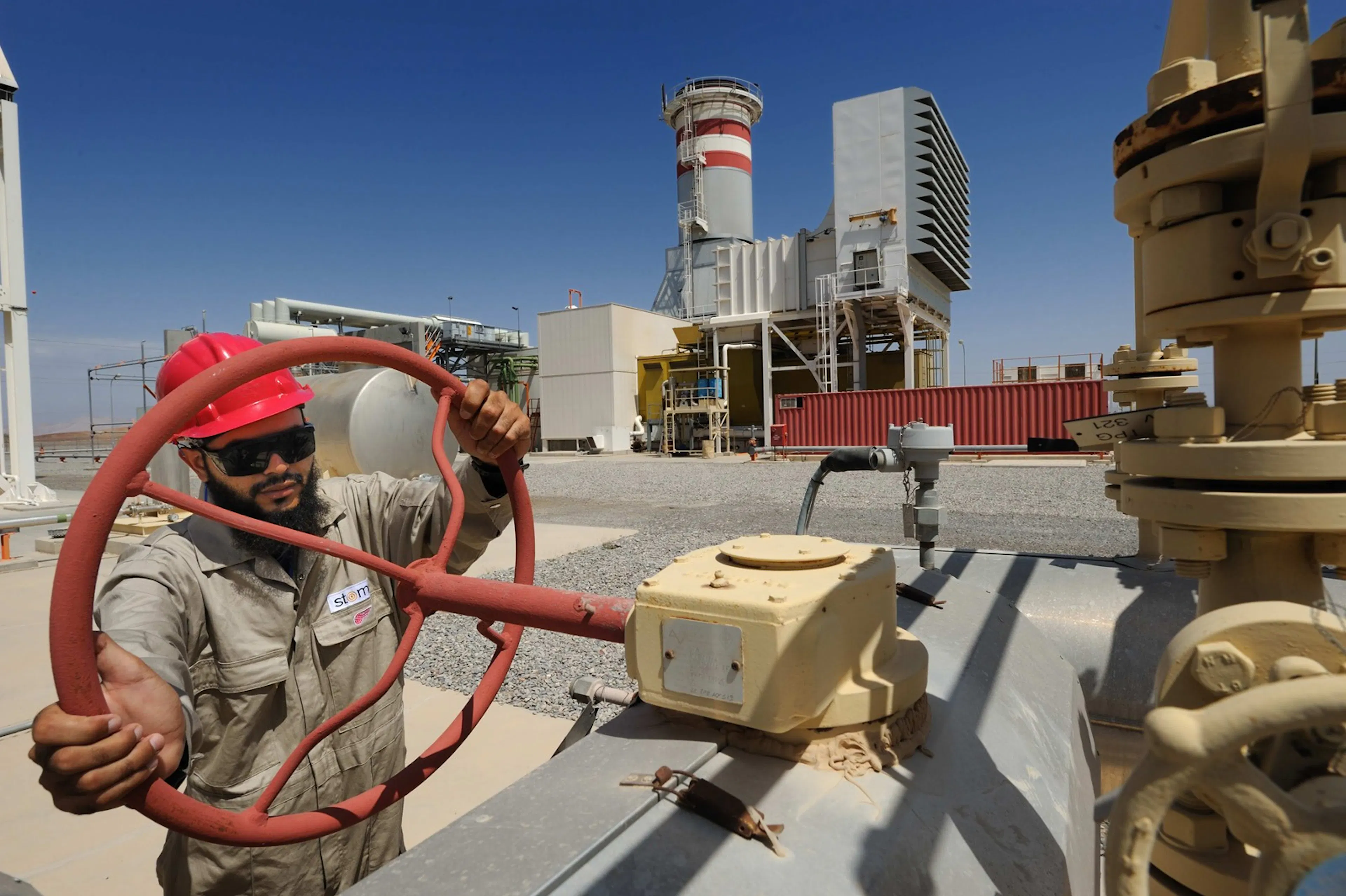 A worker in a red hard hat and coveralls operating equipment at an industrial gas plant site under a clear sky