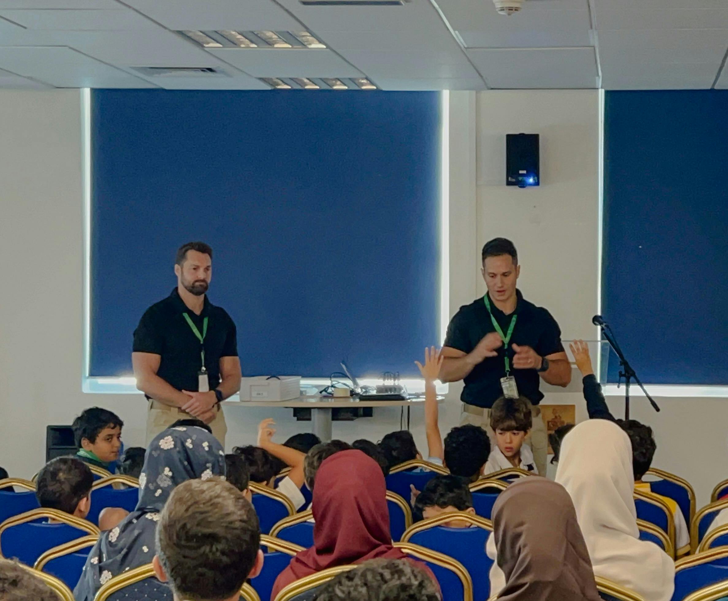 Two presenters in black shirts are speaking to a group of students seated in a classroom with blue chairs. The students, some of whom are raising their hands, appear engaged in the session. A projection screen and microphone stand are visible in the background, alongside a slide showing a group photo.