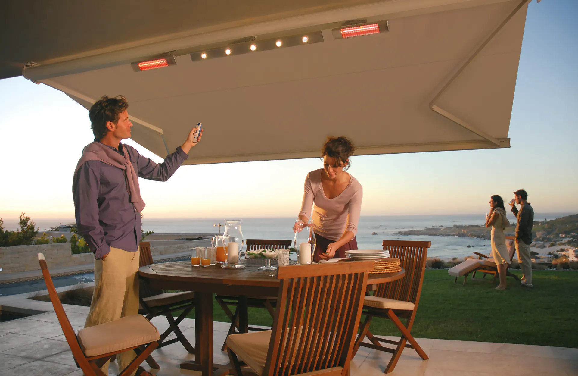 Markise med integrerte varmelamper og lys; en mann bruker fjernkontroll, en kvinne dekker et utendørs spisebord. Kontekst: terrasse ved kysten med havutsikt i solnedgang.