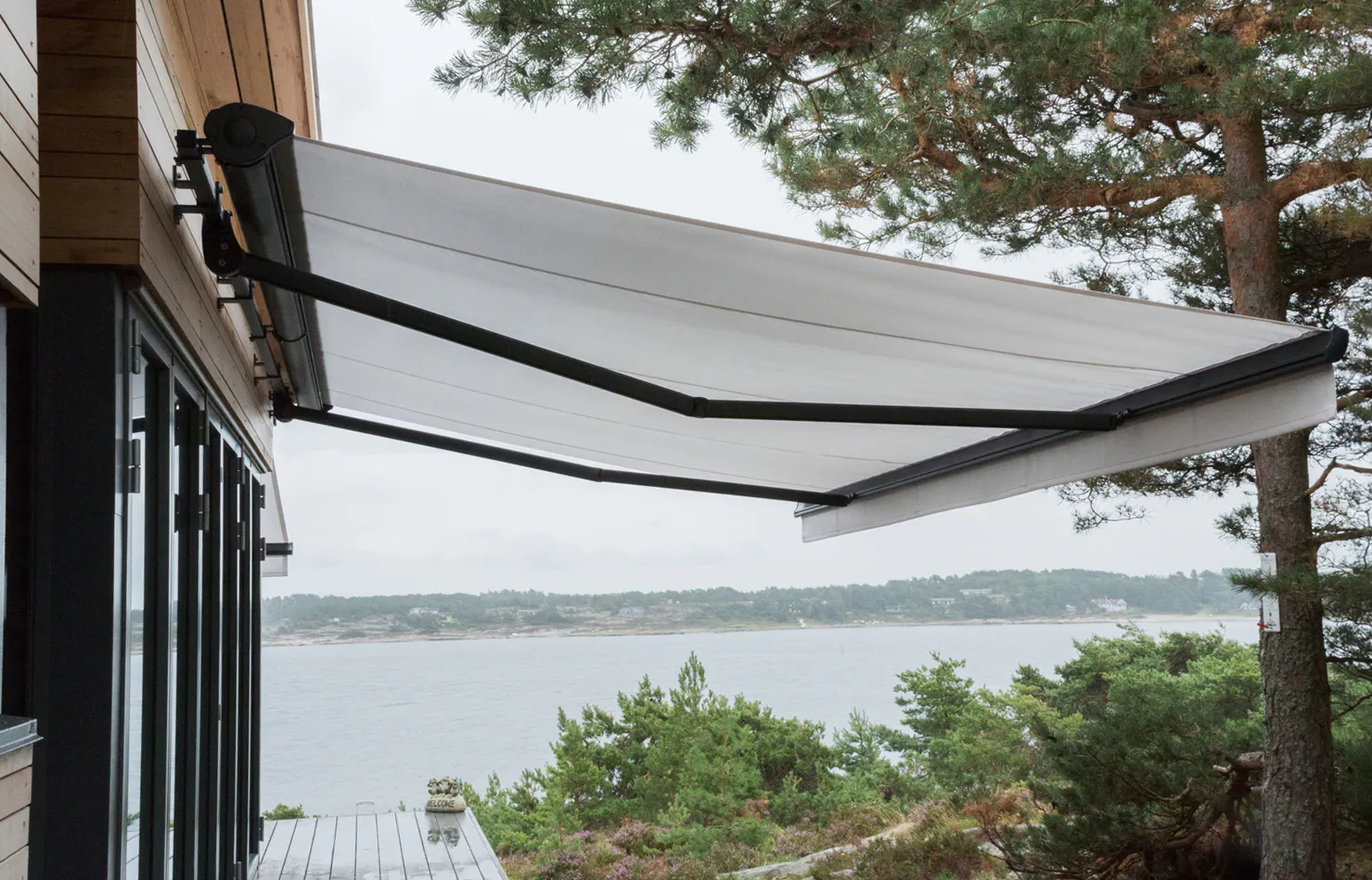 Markise utrullet over en treterrasse; veggmontert på huset til venstre og gir skygge for uteplassen. I bakgrunnen ses kystlinje med sjø, holmer og furutrær.