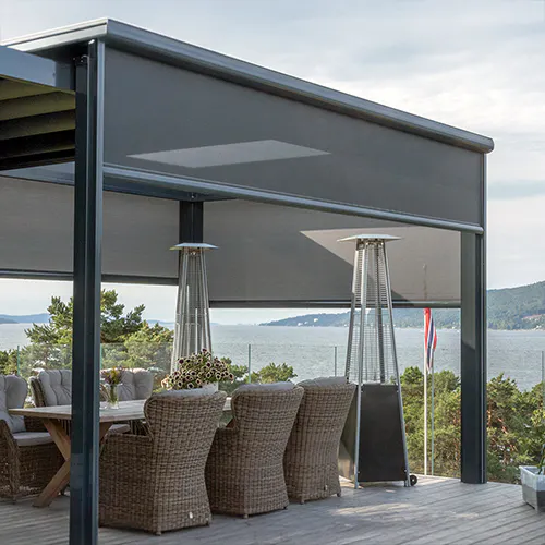 Utvendig zip-screen er nede, avskjermet mot sol og vind over en treterrasse. Under ligger et spiseområde med rottingstoler, terrassevarmere og utsikt mot fjord og furukledd kyst.