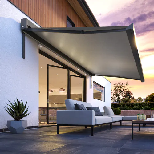 Markise montert over terrasse, utspilt og gir skygge over utesofa og lavt bord. Moderne husfasade med store glassdører, plantekrukke og kveldsfarget himmel i bakgrunnen.