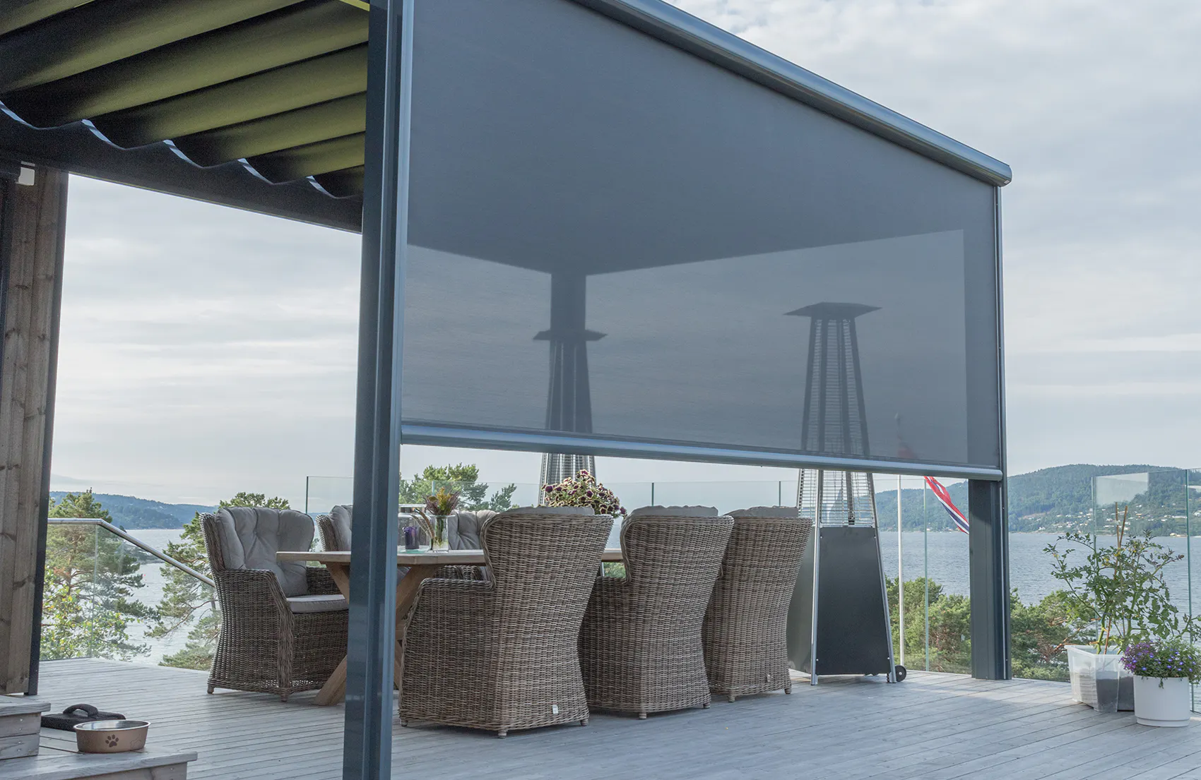 Et nedrullet, transparent zip-screen beskytter og skjermer en pergolaterrasse; omkranset av et spisebord med seks kurvstoler, terrassevarmer og utsikt mot fjord og bru under overskyet himmel.