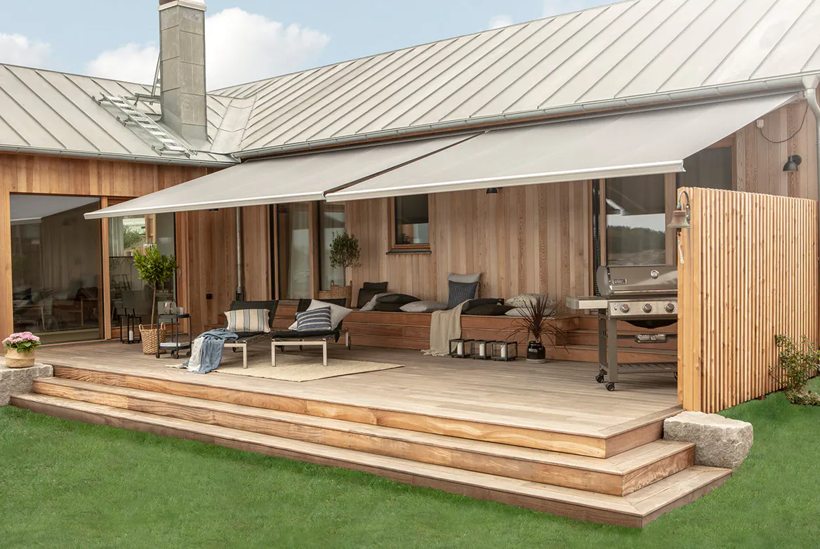 To uttrekkbare grå markiser skygger en treterrasse; de dekker en sittegruppe med sofaer, bord og puter foran en moderne trekledd bolig med gressplen og grill.