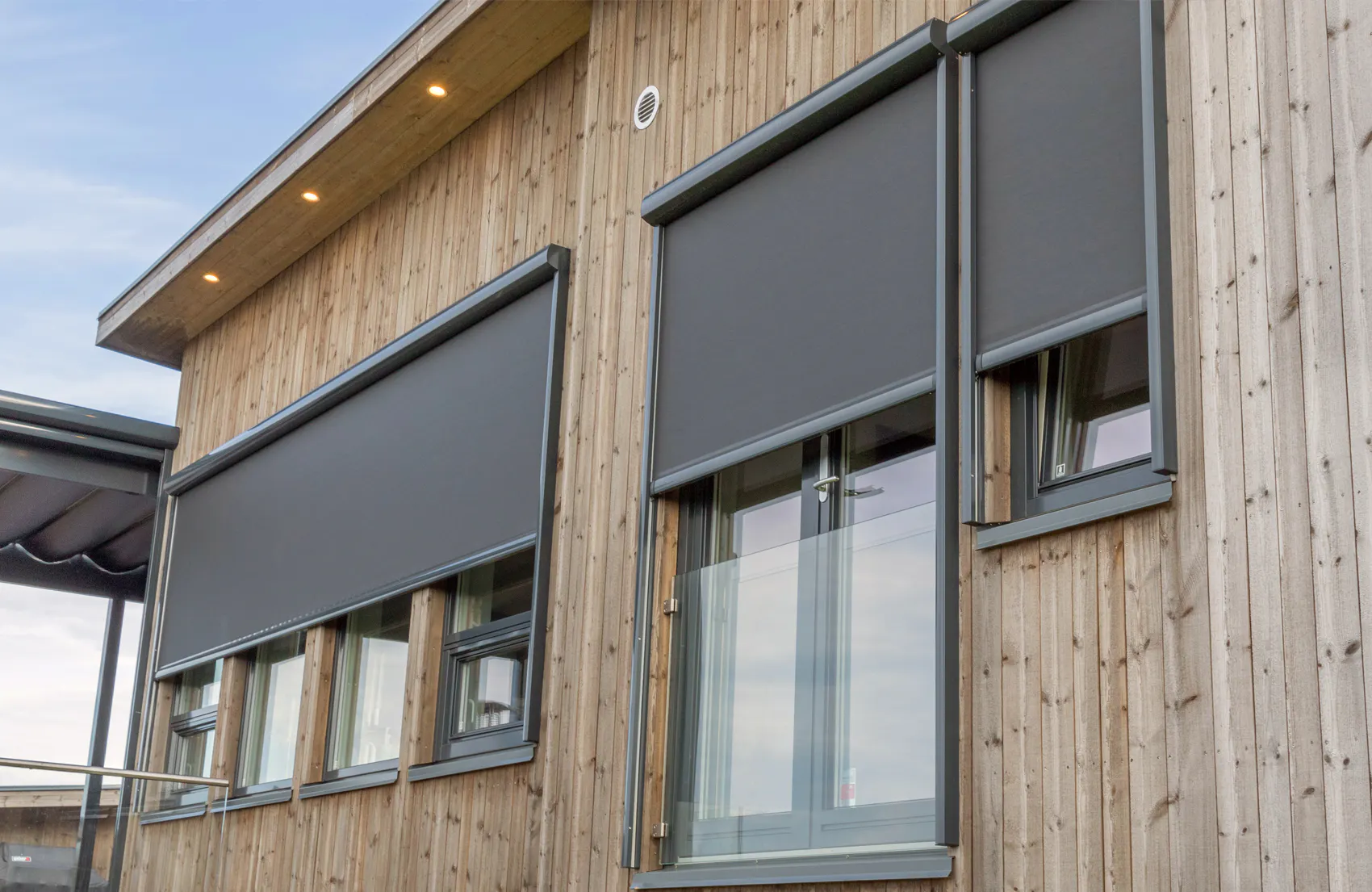 Utvendige rullegardiner (screens) er senket foran tre store vinduer og en glassdør på en moderne trefasade, og gir sol- og innsynsbeskyttelse under overskyet himmel.