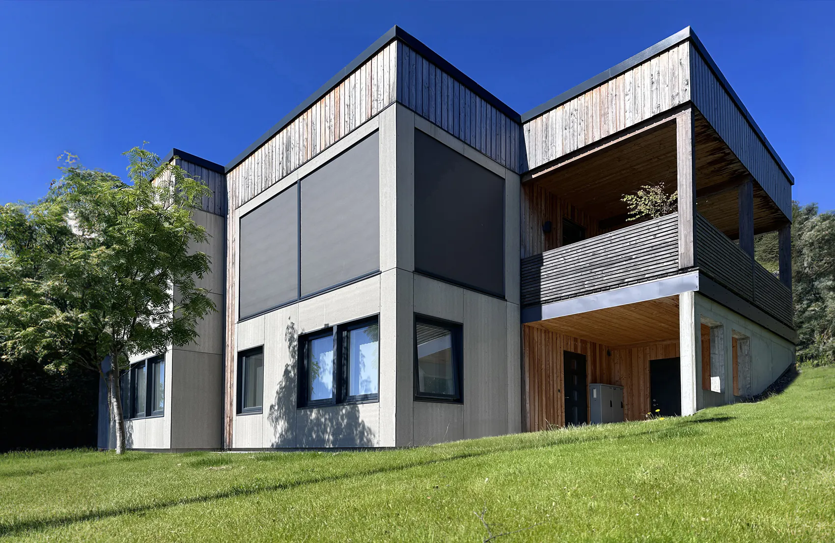 Utvendig rullegardin/solskjerming er trukket ned foran store hjørnevinduer på en moderne trekledd bolig. Solskjermingen dekker vinduene; rundt ligger grønt gress, et tre og klar blå himmel.