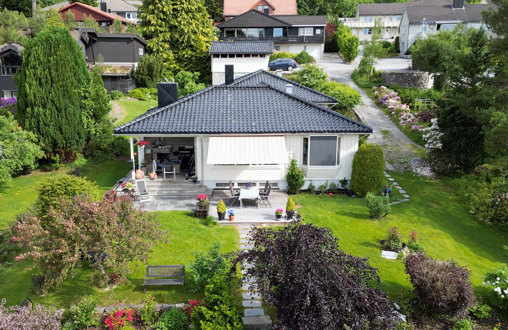 En beige markise er rullet ut over terrassevinduet og gir skygge for uteplassen. Enebolig med terrassemøbler, steinlagt platting, velstelt plen og hage i et rolig boligområde.