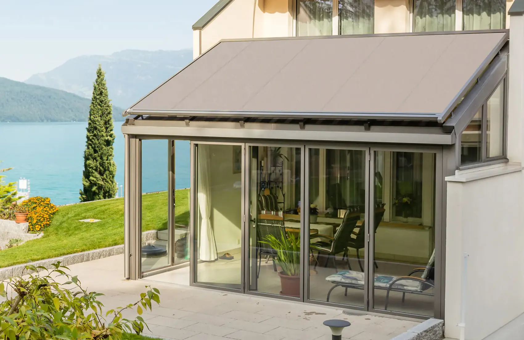 Utvendig markise er trukket ut og skygger en glassveranda med skyvedører; møbler og planter står på en terrasse ved en innsjø med fjell i bakgrunnen.