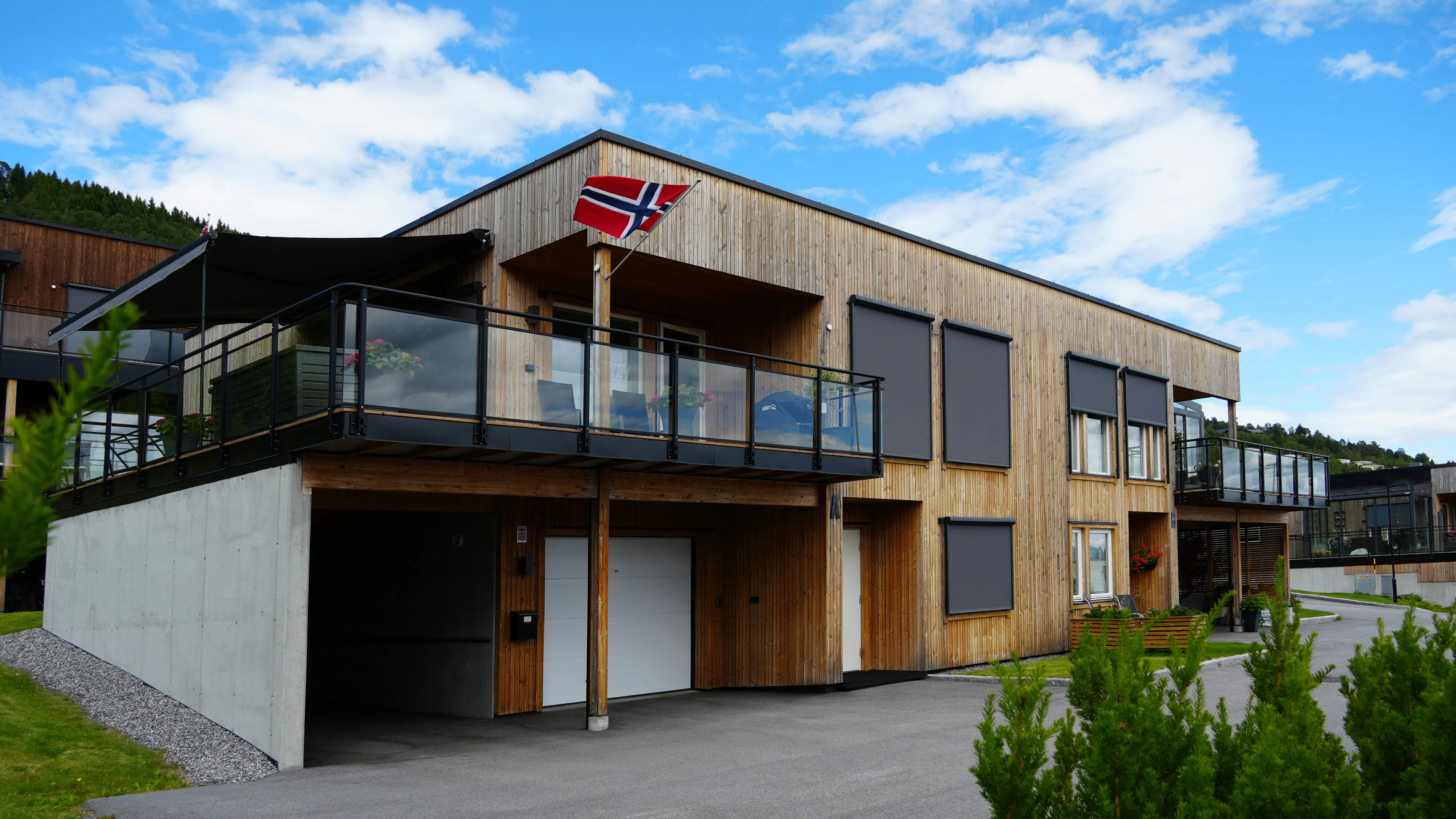 Utvendige rullegardiner/solskjermer er trukket ned foran flere vinduer på en moderne, trekledd tomannsbolig med glassbalkong og norsk flagg, ved innkjørsel og velstelt uteområde.
