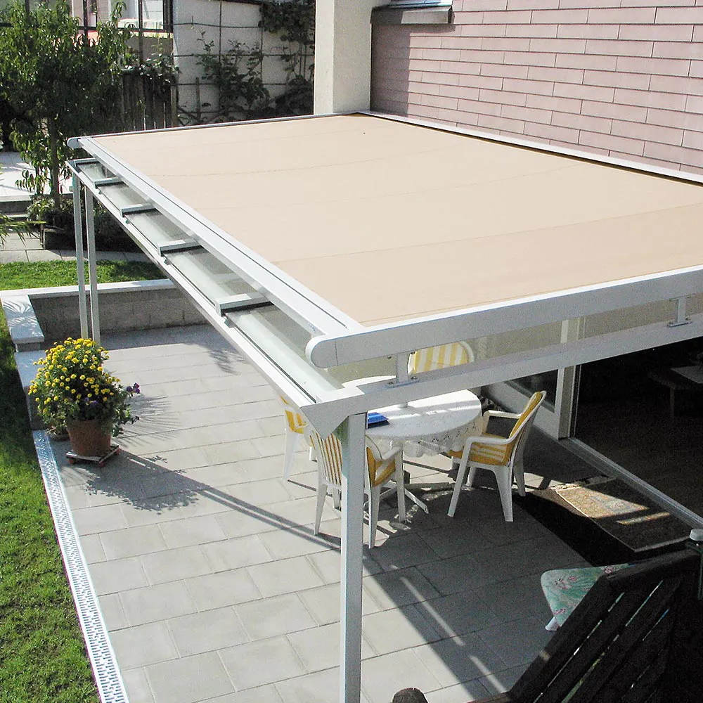 En hvit, rammefestet markise med beige duk dekker en flislagt uteplass; under står et bord med stoler, en potteblomst og husfasade i bakgrunnen.