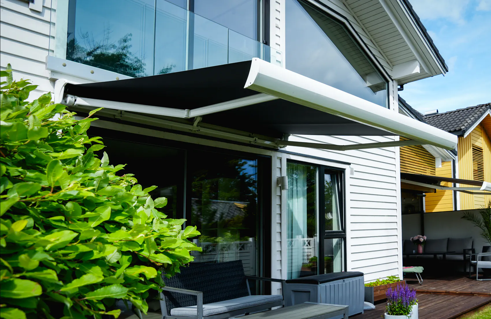 Utvendig markise er trukket ut og gir skygge over terrasse med sittegruppe, foran hvit trevilla med store glassvinduer; hekk til venstre og en gul nabobolig i bakgrunnen.
