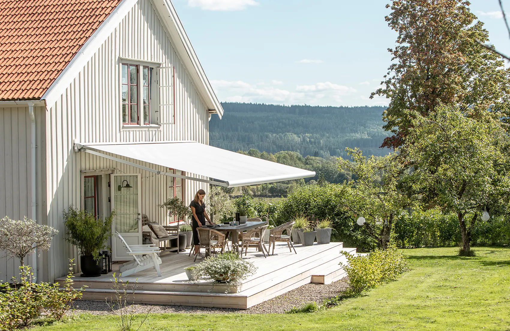 Hvit terrassemarkise er utfelt over en treplatting; en person ordner bordet under den. Hvitt trehus og hage med gress, trær og skog/innsjø i det fjerne.