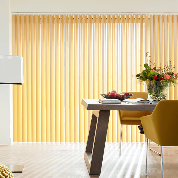 Vertikale persienner demper dagslys foran et spisebord med gul stol, fruktskål, åpen bok og en vase med blomster i et lyst, moderne rom.