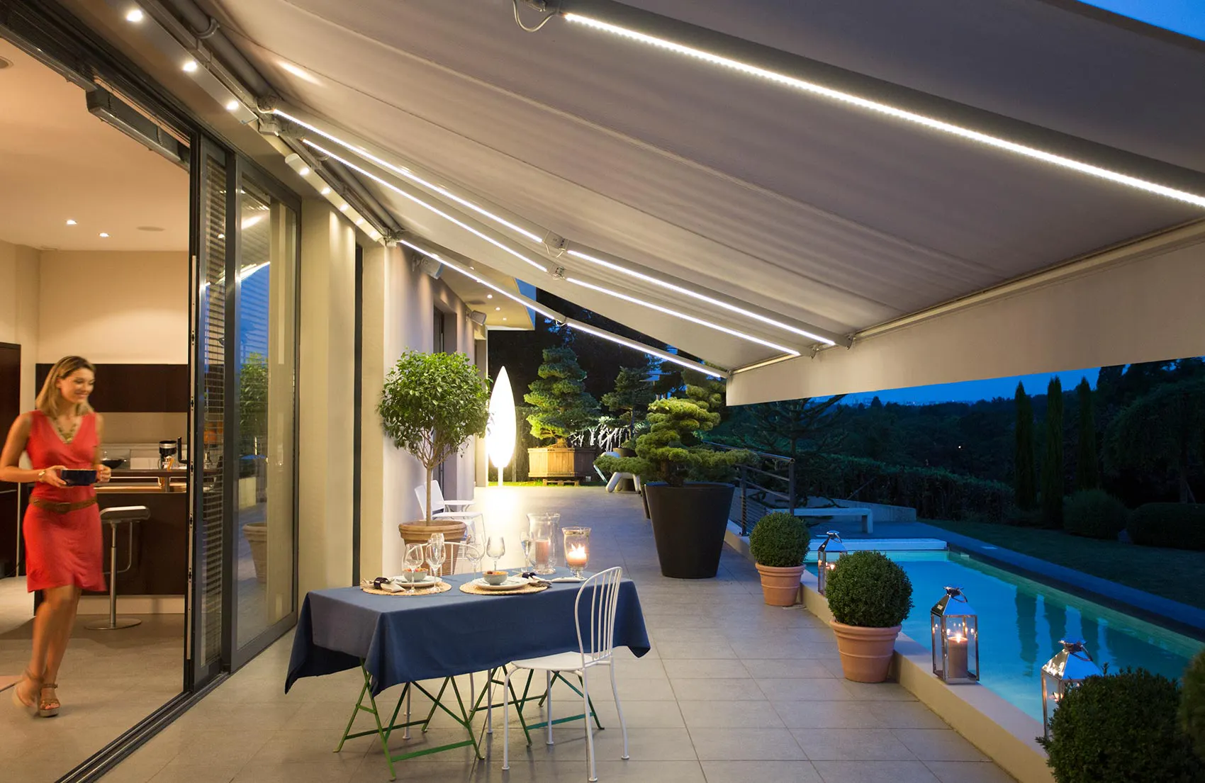 Utvendig, utbrettet terrassemarkise med integrerte LED-lys over et dekket spisebord; markisen skygger en flislagt terrasse med store plantekrukker og opplyst svømmebasseng, åpen glassdør mot kjøkken.