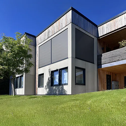 Utvendige screenmarkiser er trukket ned over to store fasadevinduer på et moderne, trekledt boligbygg; grønn plen i forgrunnen og klar blå himmel i bakgrunnen.