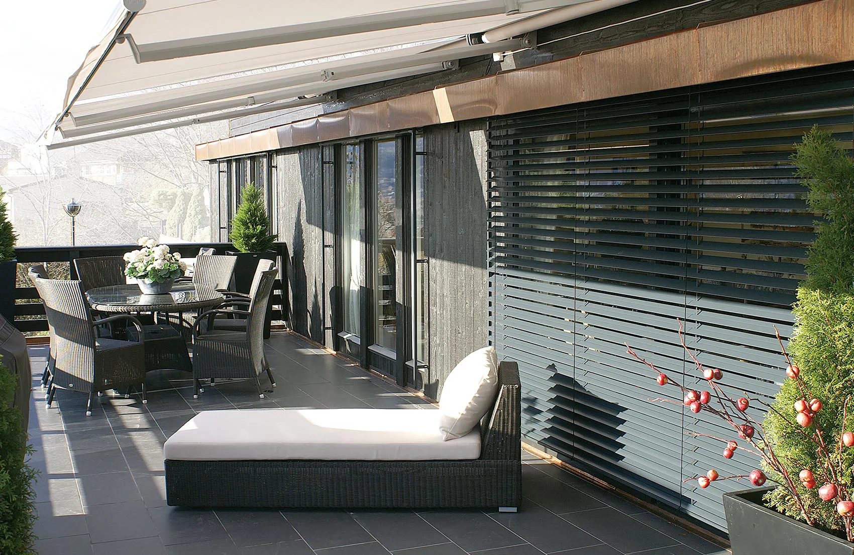 Utvendige lamellpersienner (objekt) dekker glassdører, delvis senket og justert (handling); på en solrik terrasse står loungesofa, spisebord med stoler og dekorative planter under et overbygg (kontekst).