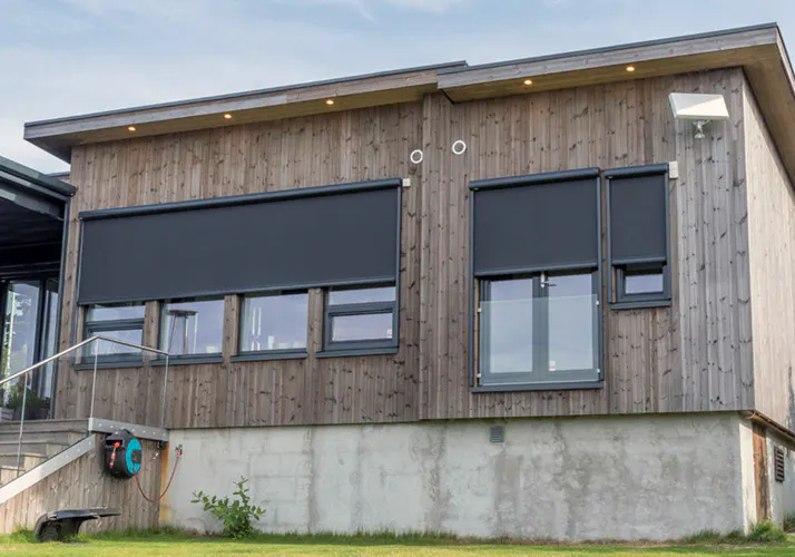 Utvendige screens dekker tre vindusfelt på en moderne trekledt fasade, nede mot betonggrunn; gressplen foran og en hageslangetrommel montert på veggen.