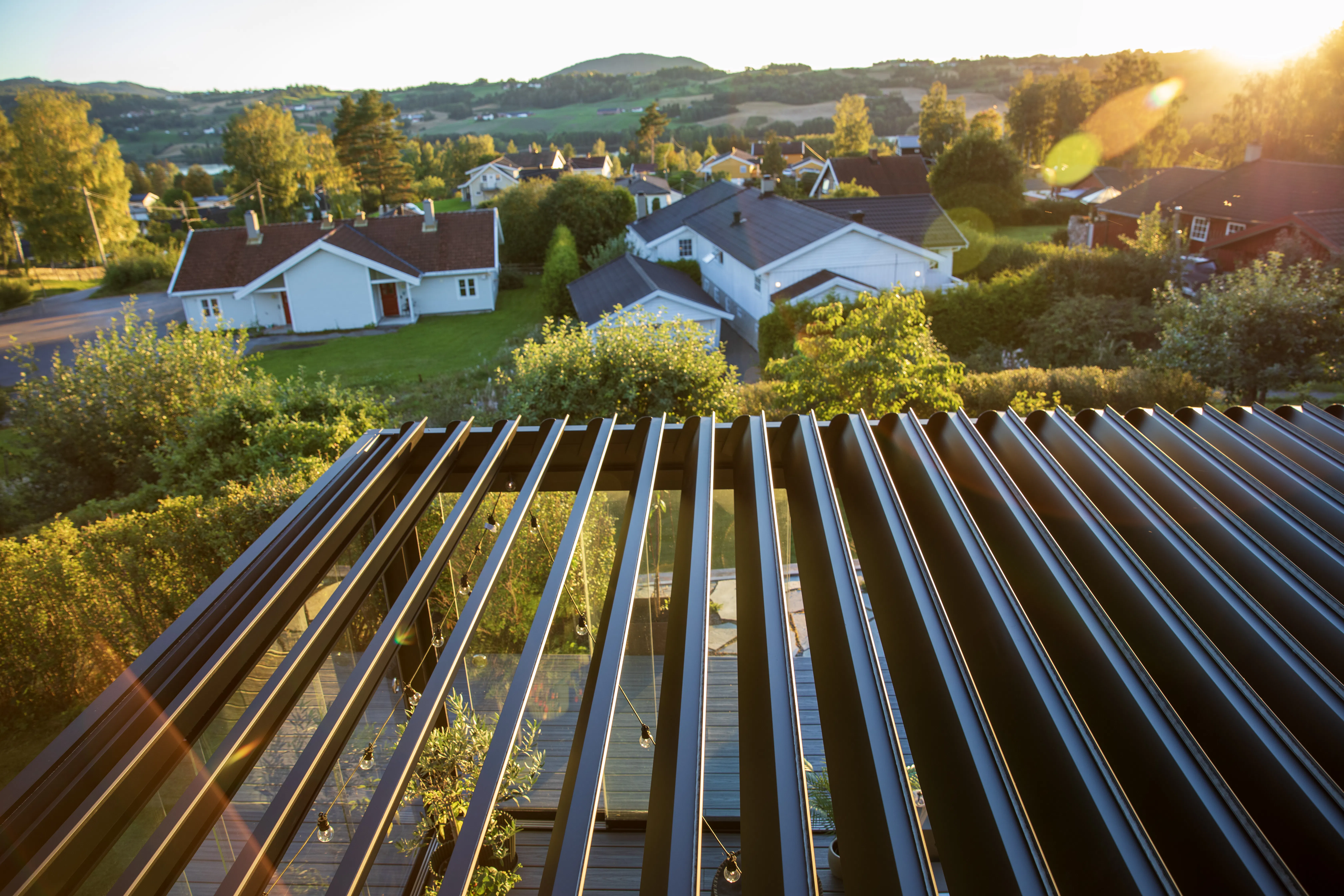 Lamelltak med mørke, parallelle lameller som gir skygge og slipper sollys; sett over terrasse med hager, hvite eneboliger og åser i kveldslys.