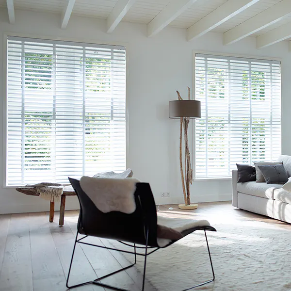 Hvite persienner på to store vinduer filtrerer dagslys inn i en lys, minimalistisk stue med hvite takbjelker, lys sofa, svart lenestol, stålampe og lyst tregulv.