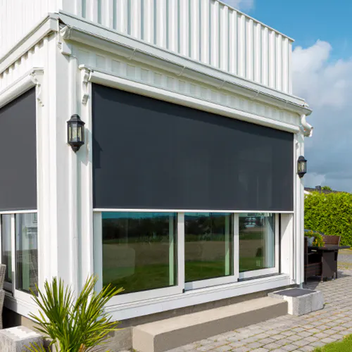 Utvendig rulleskjerm dekker store glassvinduer, gir solskjerming og privatliv på en hvit, moderne husfasade med terrasse, utelamper, plante og gressplen under blå himmel.